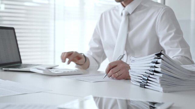 Businessman working at a desk, reviewing official documents, writing with a pen, and using a calculator, managing a stack of organized paperwork in a professional office environment. Audit and taxes
