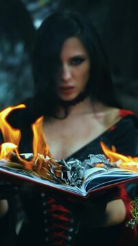 Fantasy woman sexy witch holds old burning book in hands, bright flame fire, Girl fairytale goth reads magic spell. Black red dress halloween gothic costume. Dark deep forest tree. Light on evil face
