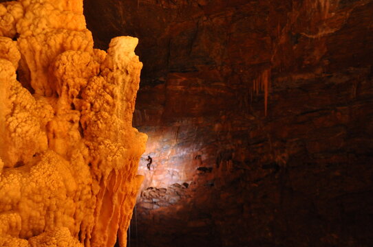 Orange calcite formation in the foreground with a tiny silhouetted figure on the distant wall of the vast Gouffre G&eacute;ant de Cabrespine cave, Aude, France.