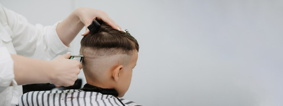 Cute Caucasian boy in a barbershop getting a haircut from a female hairdresser. space for copying. Stylist's hands with tools. Children's fashion in the salon.
