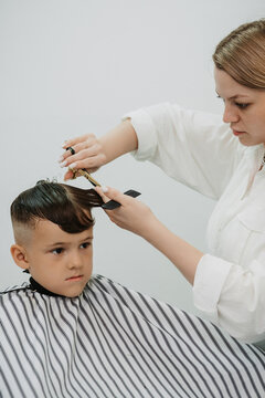 A female hairdresser cuts a little boy's hair with a comb and scissors. A professional hairdresser and a child client in a modern barbershop.
