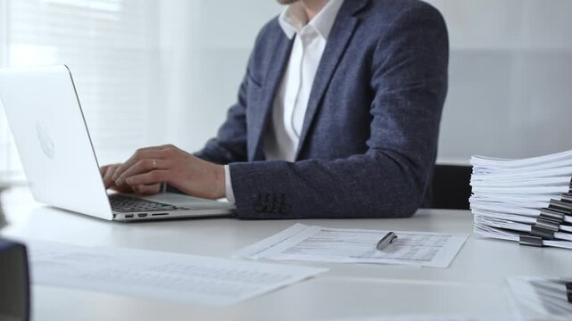 Businessman typing on laptop amid stacked documents and reports, focused on financial data and paperwork in a modern office, conveying productivity, management, and analysis. Audit and taxes