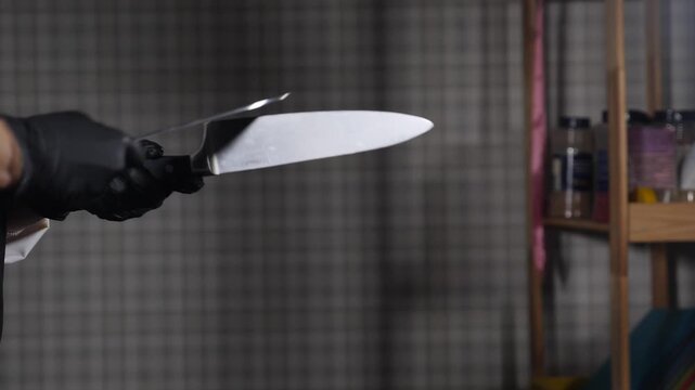 Close-up of chef in black gloves sharpening two kitchen knives against each other. Professional culinary tool maintenance, blurred spice shelf background, skilled manual work Slow motion 4k video