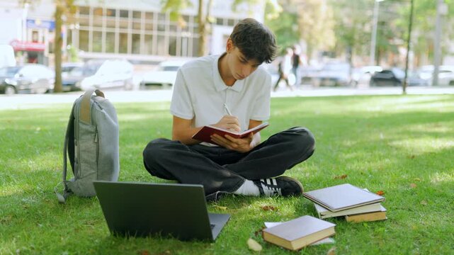 Focused young guy writing in a notebook while sitting on the park grass.