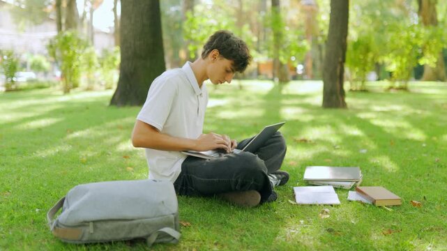 Focused young student typing on a laptop while sitting on the park grass.