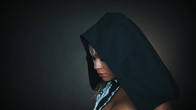 Closeup fantasy african american woman priestess looks around. Art video beauty lips headshot. Girl vampire queen looking at camera hypnotizes eyes sexy face. Gothic style Black dress hood on head 4k