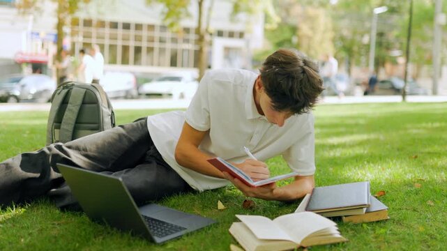 Attentive caucasian student writing notes while lying on the outdoor grass.