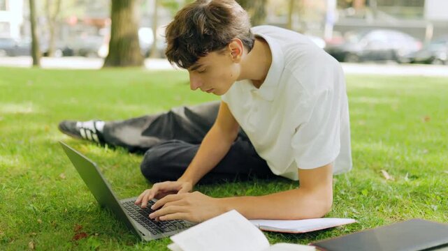 Focused male learner typing on his computer while lying on the park grass.