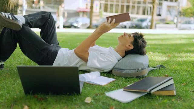 Caucasian male learner covering his face with a book on the campus grass.