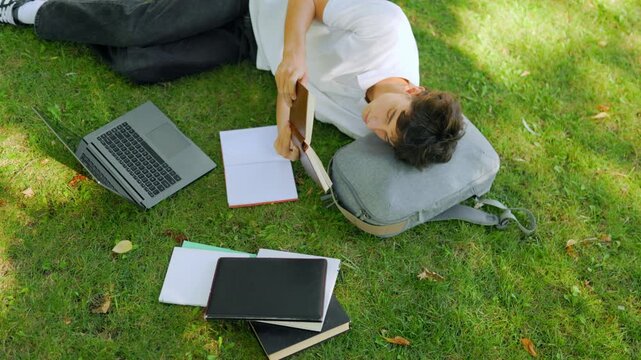 Dedicated male learner sleeping with a book on the campus grass outdoors.