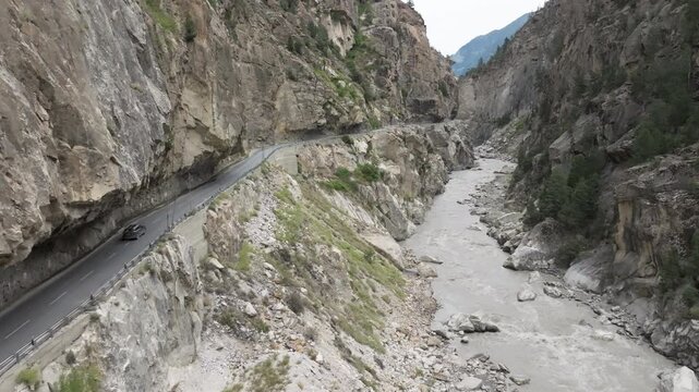 Aerial video of vehicles including a truck on a road carved into a sheer rock wall with patches of green vegetation above a grey river gorge in Kinnaur Himachal Pradesh India shot by a drone