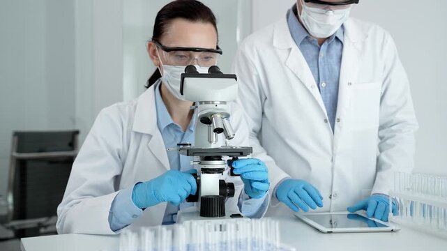 Two biotechnology professionals examining biological samples with a microscope and using a digital tablet for data entry during a scientific experiment in a life science lab
