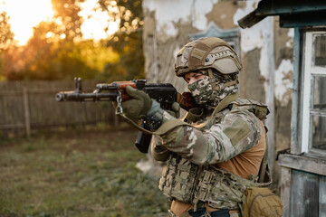 Fototapeta premium Military soldier posing at camera with assault rifle against the backdrop of an old village house
