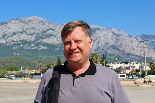 A portrait of an adult man squinting in the bright sun against a backdrop of blurred, majestic mountain peaks.