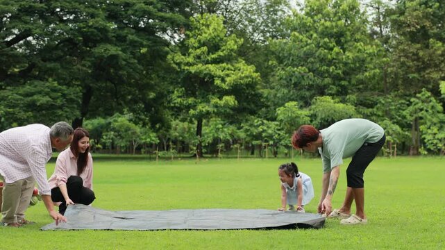 unfold picnic mat in green city park. Happy Asian family having fun time on picnic
