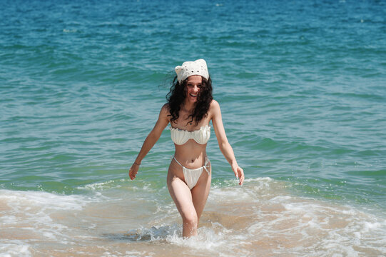a modern young woman with long curly hair in a white swimsuit on a sandy beach, walking to the sea.