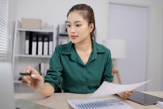Asian businesswoman working at her desk, reviewing financial reports and documents in a modern office environment.