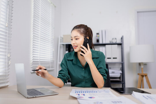 Asian businesswoman working in an office, reviewing documents while using a smartphone.