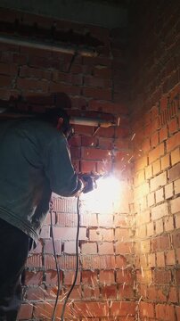 Worker in a protective mask welding a steel pipe on a brick wall at a construction site