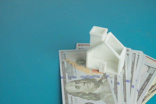White miniature house model resting on a stack of US dollar bills over a blue background with copy space. Concept of real estate investment, mortgage, property loan and saving money for a new home.
