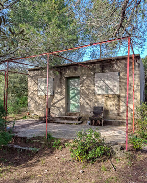 ​Abandoned concrete hut with red metal pergola frame and broken chair in pine forest. Old rural cabin with closed shutters hidden in woods on sunny day.