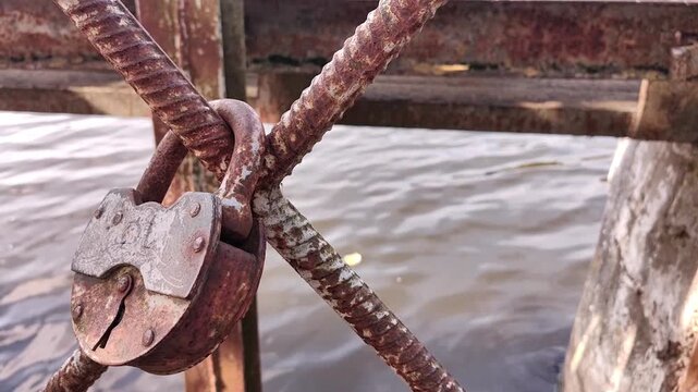 A rusty padlock hangs on a metal fence beside flowing water, weathered texture, industrial detail, river scenery, rustic landscape.