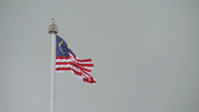 Malaysian national flag, Jalur Gemilang, waving in the wind on a tall white flagpole against a minimalist overcast gray sky, with ample copy space.