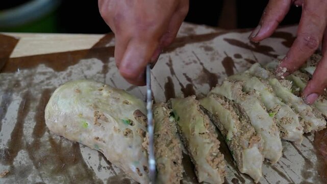 Closeup hands slicing meat stuffed dough roll with cleaver knife on cutting board, savory street food preparation.