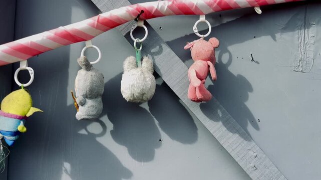 Small plush toys hanging from a candy cane pole against a gray wooden wall in bright sunlight. Playful background, cute stuffed animals, outdoor shadow, festive theme, and decorative toys.