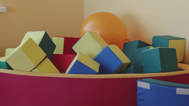 Brightly colored soft foam blocks filling a ball pit in a children's playroom. A large inflatable ball rests in the corner of the room
