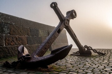 Fototapeta premium Old rusty anchors on cobblestone quay in morning fog