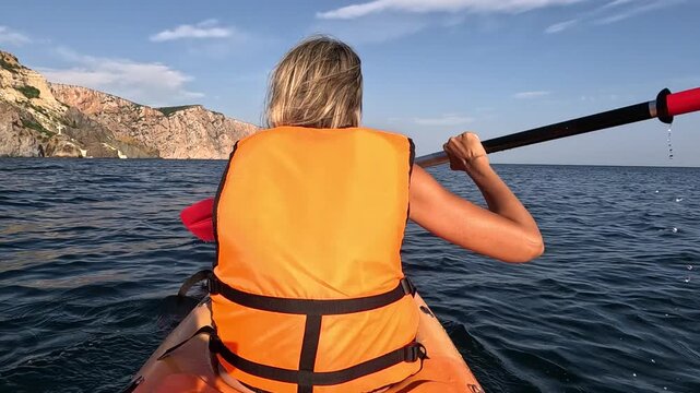 Rugged limestone cliffs rise above calm open sea as an active woman rows an orange kayak. Behind the subject angle shot.