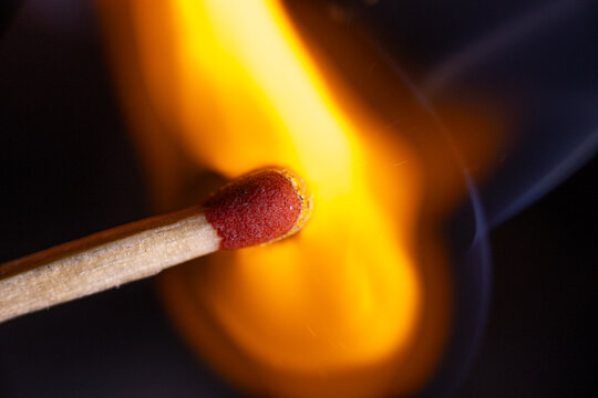 Single wooden match igniting with bright orange flame and glowing red match head against dark background in dramatic closeup macro photograph