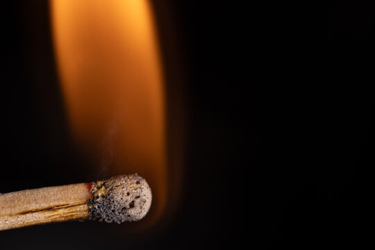 Smoldering wooden match with glowing orange flame and textured blackened head against a deep black background, dramatic closeup macro photograph of combustion