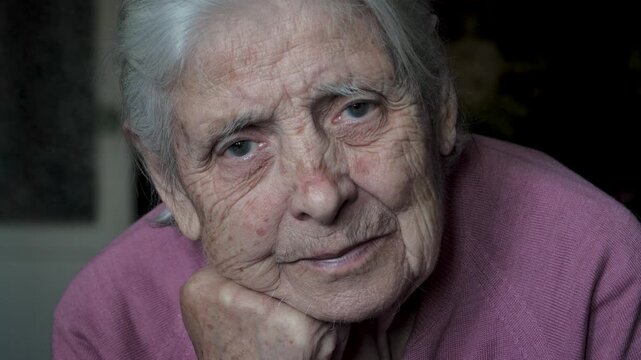 Close up portrait of old woman 80 90 years with grey hair and deep wrinkles looking into camera with kind representing wisdom experience. Woman suffers from tremors and pigmentation spots on skin.