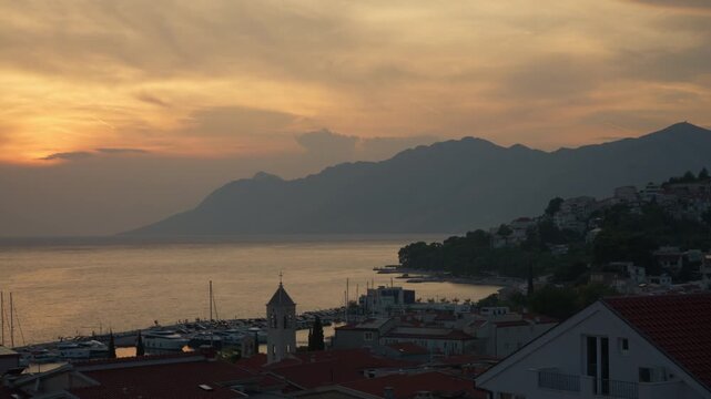 Sunset time lapse over coastal resort town of Baska Voda in Croatia. Historic church bell tower and tile roofs overlook harbor beside Adriatic sea. Scenic Mediterranean summer vacation spot.