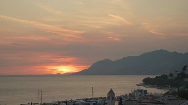 Sunset time lapse over Baska Voda resort on Adriatic coast in Croatia. Yachts rest in harbor beneath scenic mountains. Peaceful evening scene highlights coastal Mediterranean summer vacation.