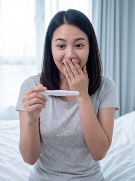 Positive pregnancy test asian woman surprised happy result bedroom fertility Surprised Asian woman covers her mouth while showing the result on her bed for reproductive health and motherhood campaigns