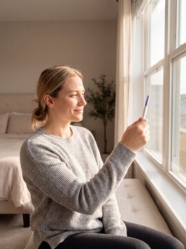 Positive pregnancy test woman window hopeful result fertility motherhood sunlight A hopeful blonde woman raises the result toward morning window light for conception and motherhood campaigns