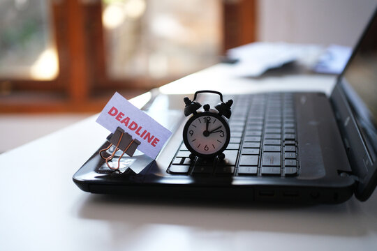 The word "Deadline" and a small alarm clock on a laptop keyboard. Concepts of deadlines and work; motivation and management; racing against time; working under pressure.