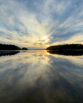 A serene sunset casts a striking golden glow through dramatic clouds, reflecting perfectly on a calm forest lake framed by dark, rocky shores.in Finland.
