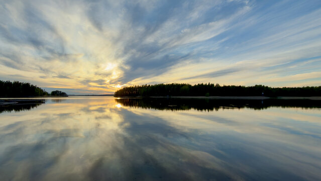 A serene sunset casts a striking golden glow through dramatic clouds, reflecting perfectly on a calm forest lake framed by dark, rocky shores.in Finland.