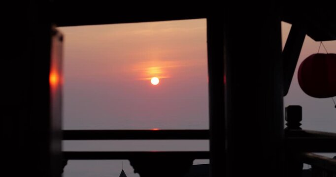 Golden morning sun rises above dense sea of clouds, viewed through dark silhouette posts of traditional wooden pavilion structure. Warm dawn light reflects off side of pillar