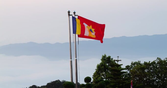 National flag of Vietnam and multi-colored Buddhist flag flutter on chrome poles during breezy morning. A vast sea of white clouds blankets valley beneath distant mountain ridges