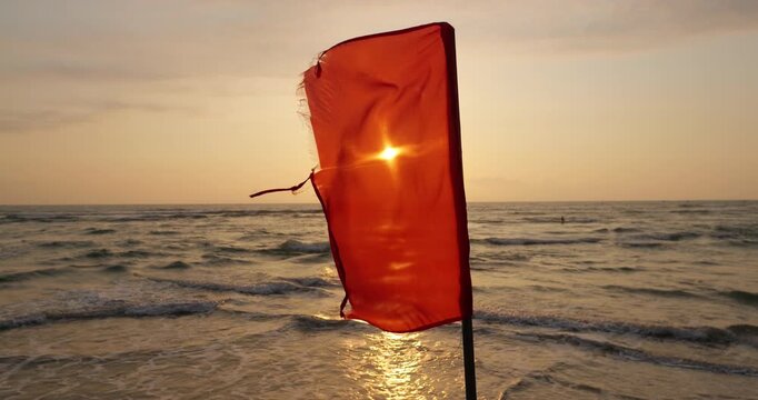 Close view of slightly tattered red warning flag fluttering in strong morning breeze on beach no-swim post. Bright morning sunlight shines from behind fabric, casting glittering reflection