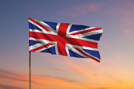 UK Great Britain national flag flying and waving in the wind, backlit in golden hour before sunset, over cloudy blue sky, symbol of British patriotism, low angle, side view