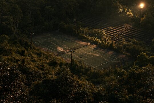 High-angle view of an abandoned tennis court engulfed by dense forest at twilight, cracked concrete surface with faded white lines barely visible beneath thick vegetation