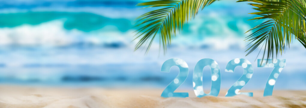 number 2027 on idyllic sand beach with palm leaf, blurred background banner with copy space for new travel season, vacation and beach holidays