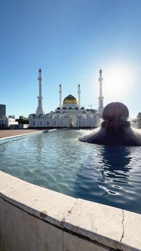 Reflecting Abu Nasr Al-Farabi Mosque across fountain water in Astana, with white domes, tall minarets, blue sky and elegant landscaped setting, shot handheld, vertical video, 4k footage