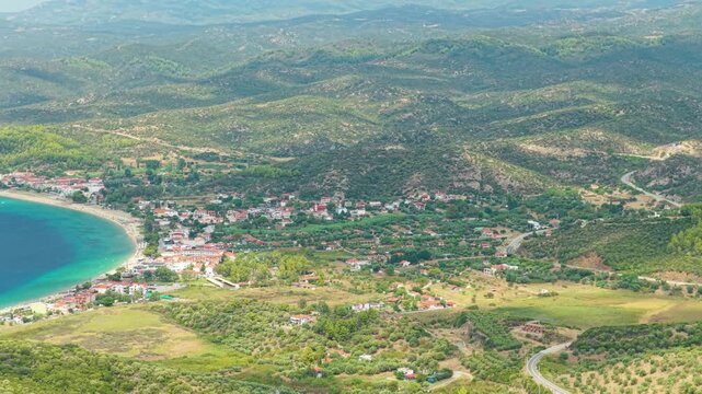 Aerial view of the coastal village of Toroni in the Halkidiki region of Greece. The turquoise waters of the bay, sandy beach, and the surrounding green hills and countryside dotted